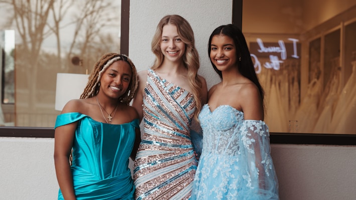 Three women in elegant evening gowns are smiling and posing together in front of a window. One wears a turquoise off-shoulder dress, the second wears a sequined gown with diagonal stripes, and the third wears a light blue dress with floral lace details. The background shows an indoor setting with a hint of wedding dresses and a sign.