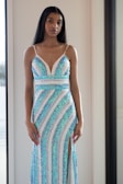 A woman stands indoors wearing a shimmering dress with blue and white sequins arranged in a striped pattern, including a thigh-high slit. Her long black hair is straightened, and she has a calm expression on her face.