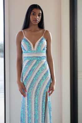 A woman stands indoors wearing a shimmering dress with blue and white sequins arranged in a striped pattern, including a thigh-high slit. Her long black hair is straightened, and she has a calm expression on her face.