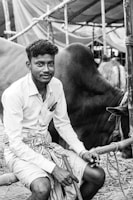 A man is seated next to a large animal, possibly a cow or bull, under a makeshift shelter made of bamboo and cloth. The setting appears rural, with informal structures and straw on the ground. The man is wearing a long sleeve shirt and a traditional cloth around his waist. He holds a stick and has a relaxed expression, with his hair neatly styled.