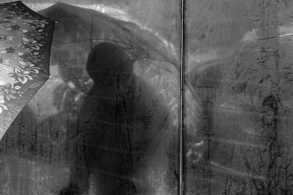 A black and white image featuring the silhouette of a person holding an umbrella against a glass surface. The surface is wet, likely from rain, and the texture of raindrops can be seen running down the glass. The umbrella has a floral pattern visible on the left.