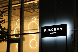 A brightly illuminated sign of a coffee shop reading 'FULCRUM COFFEE' is mounted on a building with large windows. Inside the building, circular neon lights are visible, creating a warm and inviting ambiance. The exterior has a glass facade, and there are faint reflections on the glass.