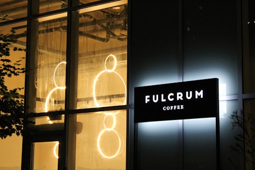 A brightly illuminated sign of a coffee shop reading 'FULCRUM COFFEE' is mounted on a building with large windows. Inside the building, circular neon lights are visible, creating a warm and inviting ambiance. The exterior has a glass facade, and there are faint reflections on the glass.