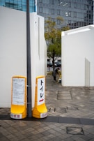 Two yellow signs are placed on a tiled pathway, pointing towards a toilet. The signs contain text and arrows directing to the right. There are white buildings on either side of the pathway, one of which has a person bending down near the entrance. A tree with green and autumn-colored leaves stands in the middle background, with modern glass-fronted buildings visible further behind.