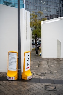 Two yellow signs are placed on a tiled pathway, pointing towards a toilet. The signs contain text and arrows directing to the right. There are white buildings on either side of the pathway, one of which has a person bending down near the entrance. A tree with green and autumn-colored leaves stands in the middle background, with modern glass-fronted buildings visible further behind.