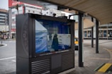 An outdoor digital signboard reflects the surrounding urban environment, including buildings and a street. The area is characterized by modern architecture and a curved wooden roof structure, with a few people and a yellow taxi visible in the background.