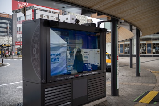 An outdoor digital signboard reflects the surrounding urban environment, including buildings and a street. The area is characterized by modern architecture and a curved wooden roof structure, with a few people and a yellow taxi visible in the background.