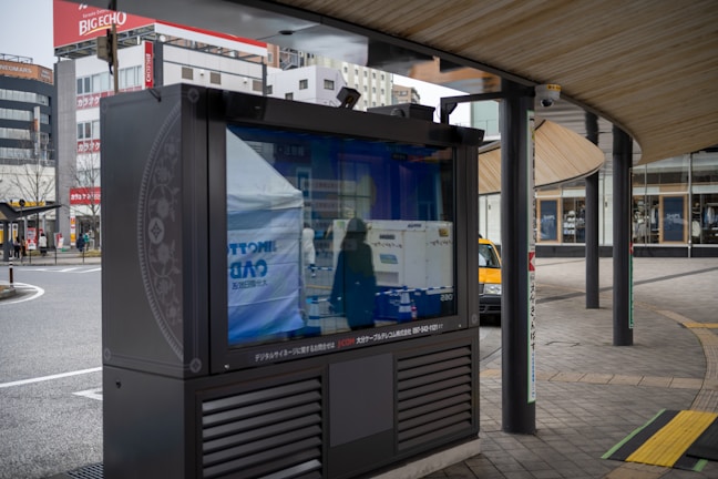 An outdoor digital signboard reflects the surrounding urban environment, including buildings and a street. The area is characterized by modern architecture and a curved wooden roof structure, with a few people and a yellow taxi visible in the background.