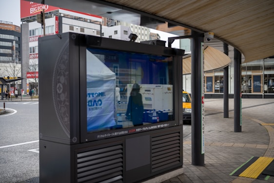 An outdoor digital signboard reflects the surrounding urban environment, including buildings and a street. The area is characterized by modern architecture and a curved wooden roof structure, with a few people and a yellow taxi visible in the background.