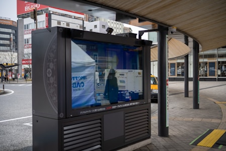 An outdoor digital signboard reflects the surrounding urban environment, including buildings and a street. The area is characterized by modern architecture and a curved wooden roof structure, with a few people and a yellow taxi visible in the background.