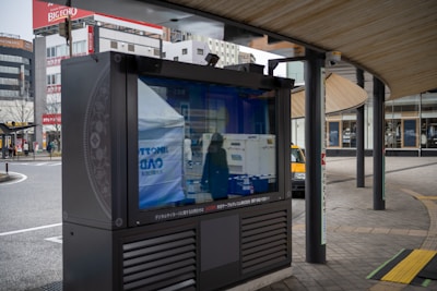 An outdoor digital signboard reflects the surrounding urban environment, including buildings and a street. The area is characterized by modern architecture and a curved wooden roof structure, with a few people and a yellow taxi visible in the background.