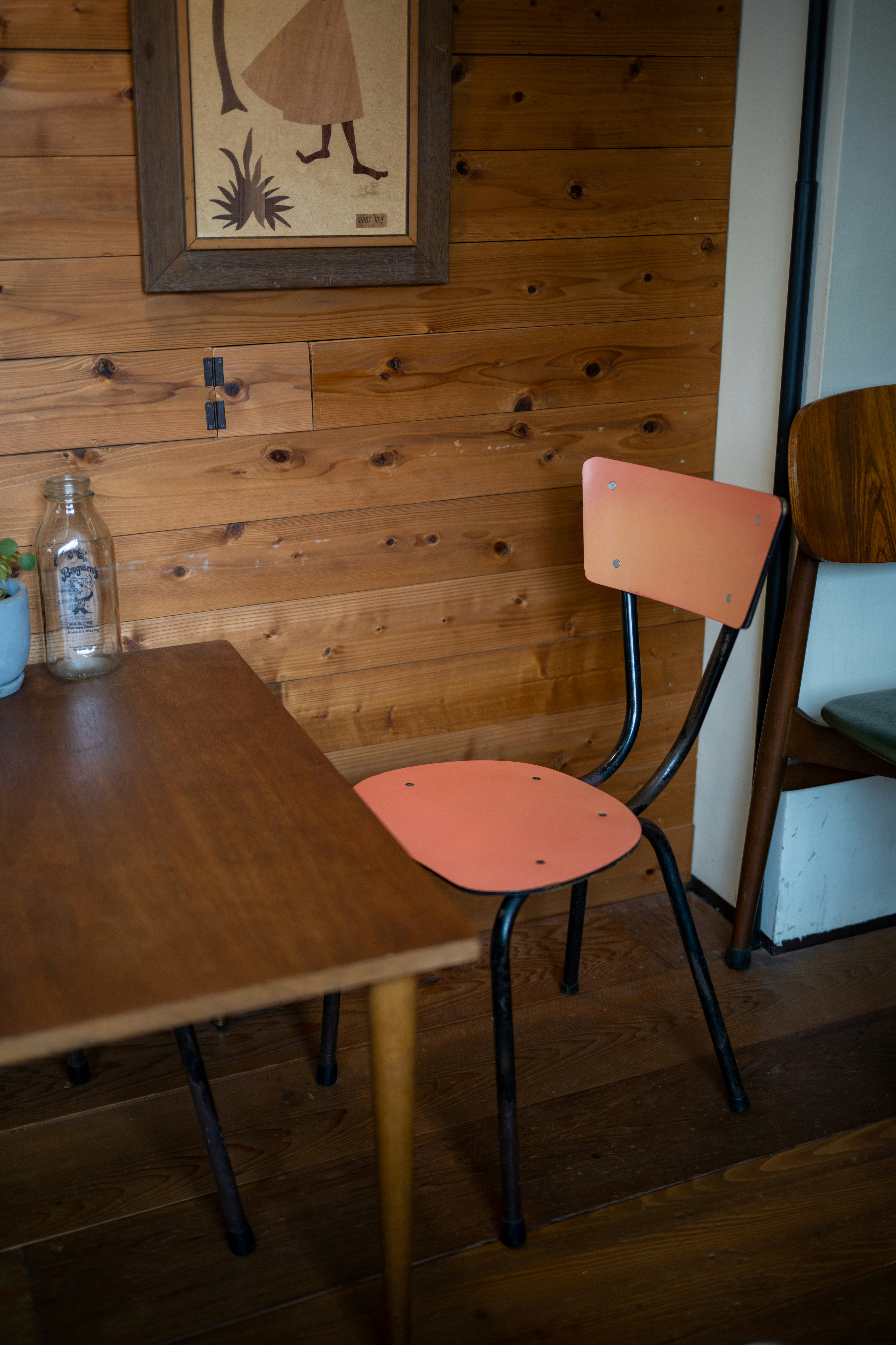 A wooden wall with a framed artwork hangs next to a corner featuring a table and two chairs. One chair has a pink-orange seat with black metal legs. A small plant and a clear bottle are placed on the table.