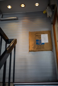 A staircase with metal handrails leads upwards alongside a corrugated metal wall. A burlap coffee sack is mounted on the wall, displaying labels indicating Costa Rican origin and details such as crop year and weight. Warm ceiling lights illuminate the area, creating a cozy ambiance.