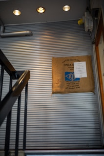 A staircase with metal handrails leads upwards alongside a corrugated metal wall. A burlap coffee sack is mounted on the wall, displaying labels indicating Costa Rican origin and details such as crop year and weight. Warm ceiling lights illuminate the area, creating a cozy ambiance.
