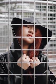 A person with red hair, wearing a black hooded jacket, is standing behind a metal grid structure with eyes closed. The expression appears calm or introspective. The grid creates a pattern over the person, enhancing the impression of separation or confinement.