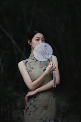 A person in traditional attire holds an ornate hand fan partially covering their face, standing against a dark, lush background of foliage. The attire has floral patterns and soft colors, and the fan displays delicate artwork.
