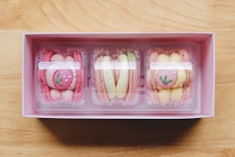 A pink box contains three compartments, each holding creatively decorated macarons. The macarons in the left and right compartments have pink shells with a design resembling a strawberry with green leaves. The middle compartment holds macarons with pale yellow and pink shells. The box sits on a wooden surface.
