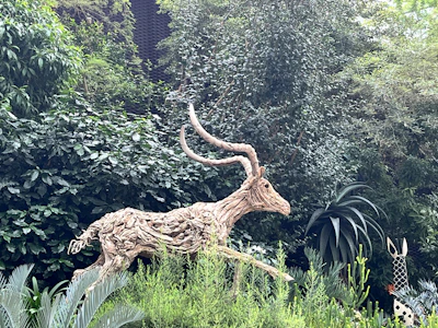 A sculpture of a deer made from twisted wooden branches, situated in a lush green garden setting. Surrounding the sculpture are dense green foliage and various types of plants, including tall leafy shrubs.
