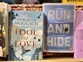 Two books are displayed side by side on a wooden surface. The book on the left, titled 'Fool for Love' by Deborah Moggach, features a cover with illustrated birds flying over a building set against a sky-blue and white striped background. The book on the right, titled 'Run and Hide' by Pankaj Mishra, has a blue cover with large white letters and an image of a small figure walking towards snowy mountains.