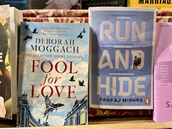 Two books are displayed side by side on a wooden surface. The book on the left, titled 'Fool for Love' by Deborah Moggach, features a cover with illustrated birds flying over a building set against a sky-blue and white striped background. The book on the right, titled 'Run and Hide' by Pankaj Mishra, has a blue cover with large white letters and an image of a small figure walking towards snowy mountains.