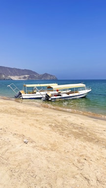 Two boats with yellow canopies are parked on a sandy beach next to a calm blue sea. In the background, rugged hills can be seen under a clear blue sky.