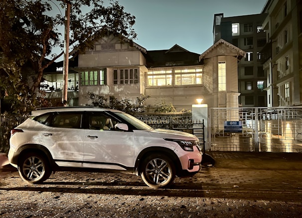 A white SUV is parked on a cobblestone street beside a large, well-lit building with classic architecture. The scene is set at dusk or nighttime, with soft lighting casting shadows from nearby trees onto the building. A sign on the gate reads 'Apollo Clinic'. Surrounding buildings are visible with windows illuminated.