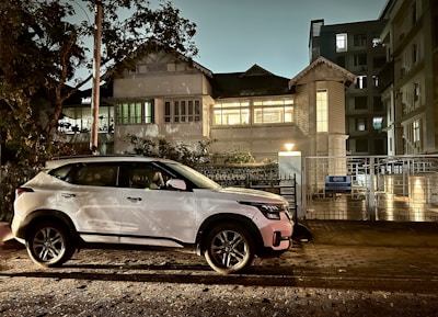 A white SUV is parked on a cobblestone street beside a large, well-lit building with classic architecture. The scene is set at dusk or nighttime, with soft lighting casting shadows from nearby trees onto the building. A sign on the gate reads 'Apollo Clinic'. Surrounding buildings are visible with windows illuminated.