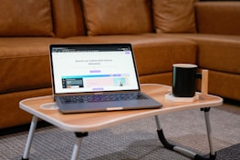 A laptop is placed on a small wooden table with folding legs, featuring a webpage about Canva Websites on its screen. The table also holds a black mug set on a white coaster. In the background, there is a brown leather sofa, and the room has a cozy and casual ambiance.