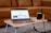A laptop is placed on a small wooden table with folding legs, featuring a webpage about Canva Websites on its screen. The table also holds a black mug set on a white coaster. In the background, there is a brown leather sofa, and the room has a cozy and casual ambiance.