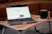 A laptop displaying a webpage is placed on a small wooden table, accompanied by a black mug on a coaster. The background consists of a brown sofa, creating a cozy indoor setting.