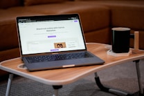 A laptop displaying a webpage is placed on a small wooden table, accompanied by a black mug on a coaster. The background consists of a brown sofa, creating a cozy indoor setting.
