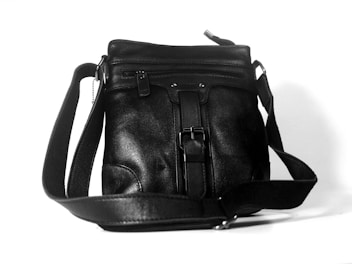 A black leather shoulder bag with a strap and buckle detail. It has a zippered pocket on the front and a smooth texture.