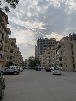 A residential area with multi-story apartment buildings lining the sides of a street. Parked cars are visible along the road, and the sky is partly cloudy with patches of blue peeking through. The buildings feature balconies and appear to be modern in design.