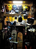 A workshop area filled with various biking and sports equipment. Several bike helmets are neatly arranged on an upper shelf. Below, there are bicycle parts, tools, and other gear. Two snowboards with graphics are leaning against the wall, and biking gear such as gloves and hats are scattered around. The area is lit with a bright overhead light, highlighting the equipment.