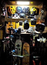 A workshop area filled with various biking and sports equipment. Several bike helmets are neatly arranged on an upper shelf. Below, there are bicycle parts, tools, and other gear. Two snowboards with graphics are leaning against the wall, and biking gear such as gloves and hats are scattered around. The area is lit with a bright overhead light, highlighting the equipment.