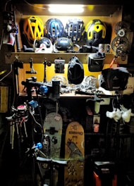 A workshop area filled with various biking and sports equipment. Several bike helmets are neatly arranged on an upper shelf. Below, there are bicycle parts, tools, and other gear. Two snowboards with graphics are leaning against the wall, and biking gear such as gloves and hats are scattered around. The area is lit with a bright overhead light, highlighting the equipment.