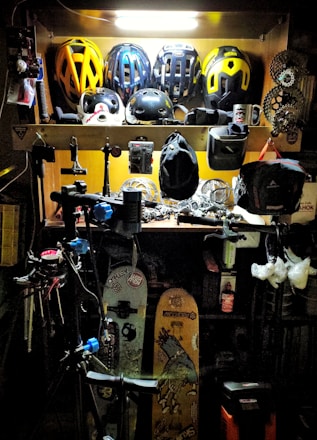 A workshop area filled with various biking and sports equipment. Several bike helmets are neatly arranged on an upper shelf. Below, there are bicycle parts, tools, and other gear. Two snowboards with graphics are leaning against the wall, and biking gear such as gloves and hats are scattered around. The area is lit with a bright overhead light, highlighting the equipment.