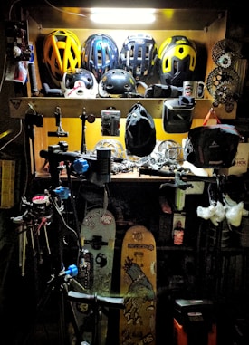 A workshop area filled with various biking and sports equipment. Several bike helmets are neatly arranged on an upper shelf. Below, there are bicycle parts, tools, and other gear. Two snowboards with graphics are leaning against the wall, and biking gear such as gloves and hats are scattered around. The area is lit with a bright overhead light, highlighting the equipment.