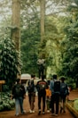A group of six people with backpacks walk along a brick path surrounded by lush greenery, heading towards a statue of a robed figure. Tall trees and dense foliage create a peaceful, natural setting.