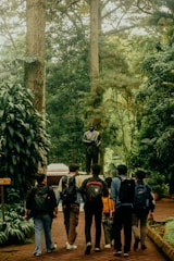 A group of six people with backpacks walk along a brick path surrounded by lush greenery, heading towards a statue of a robed figure. Tall trees and dense foliage create a peaceful, natural setting.