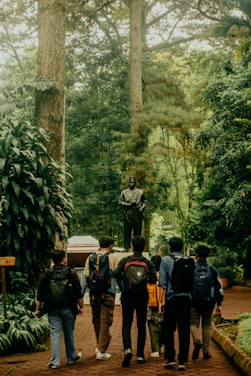 A group of six people with backpacks walk along a brick path surrounded by lush greenery, heading towards a statue of a robed figure. Tall trees and dense foliage create a peaceful, natural setting.