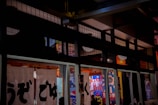 Traditional Japanese architecture with white noren curtains featuring black kanji characters. The interior has colorful posters and lanterns, creating a vibrant and cultural ambiance.
