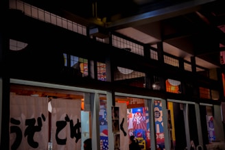 Traditional Japanese architecture with white noren curtains featuring black kanji characters. The interior has colorful posters and lanterns, creating a vibrant and cultural ambiance.