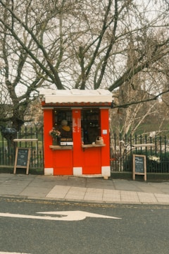 A small, vibrant red kiosk is situated on a city sidewalk, surrounded by leafless trees and a black iron fence. The kiosk has two service windows, one of which is adorned with a small wreath. Blackboards with chalk writing stand on either side, presumably displaying a menu and specials. The area is quiet, with no visible foot traffic.