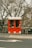A small, vibrant red kiosk is situated on a city sidewalk, surrounded by leafless trees and a black iron fence. The kiosk has two service windows, one of which is adorned with a small wreath. Blackboards with chalk writing stand on either side, presumably displaying a menu and specials. The area is quiet, with no visible foot traffic.