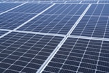 A close-up view of a large array of solar panels, with a uniform pattern of dark blue photovoltaic cells arranged in a grid. The panels are lined with silver frames, reflecting sunlight and indicating a clear day.