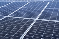 A close-up view of a large array of solar panels, with a uniform pattern of dark blue photovoltaic cells arranged in a grid. The panels are lined with silver frames, reflecting sunlight and indicating a clear day.