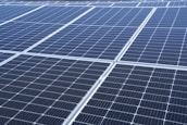 A close-up view of a large array of solar panels, with a uniform pattern of dark blue photovoltaic cells arranged in a grid. The panels are lined with silver frames, reflecting sunlight and indicating a clear day.