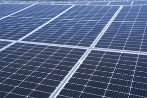 A close-up view of a large array of solar panels, with a uniform pattern of dark blue photovoltaic cells arranged in a grid. The panels are lined with silver frames, reflecting sunlight and indicating a clear day.