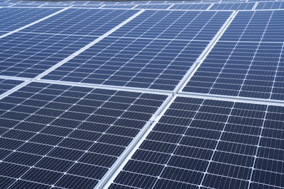 A close-up view of a large array of solar panels, with a uniform pattern of dark blue photovoltaic cells arranged in a grid. The panels are lined with silver frames, reflecting sunlight and indicating a clear day.